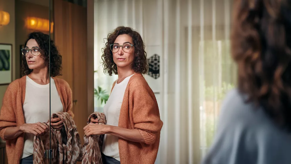 Woman with orange cardigan and glasses holding scarf in front of mirror