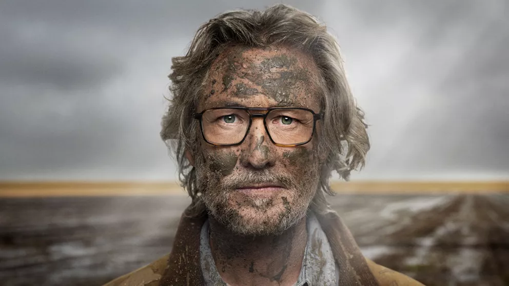 Älterer Mann steht draußen vor einem schlammigen Feld, sein Gesicht ist mit Schlamm bedeckt, aber seine Brille ist sauber