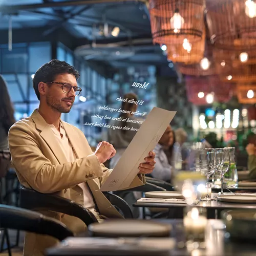 Mann im beigen Anzug trägt HOYA VisuPro-Brille zum Lesen einer Restaurant-Speisekarte