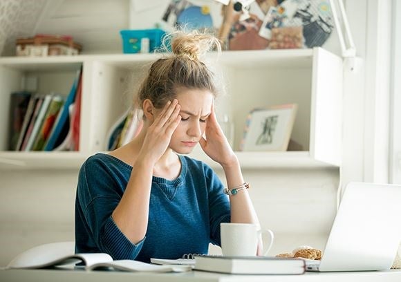 Female in a blue top sat at a desk infront of a laptop holding her head