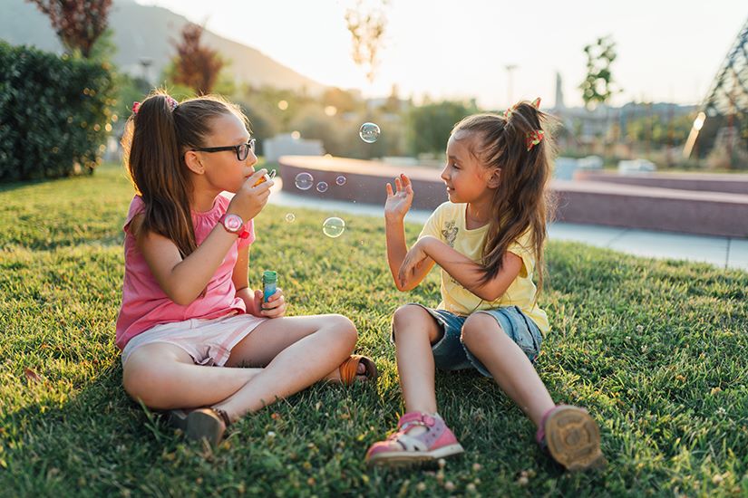 Two young girls sitting on grass playing with bubbles one wearing glasses with Hoya Vision kids lenses