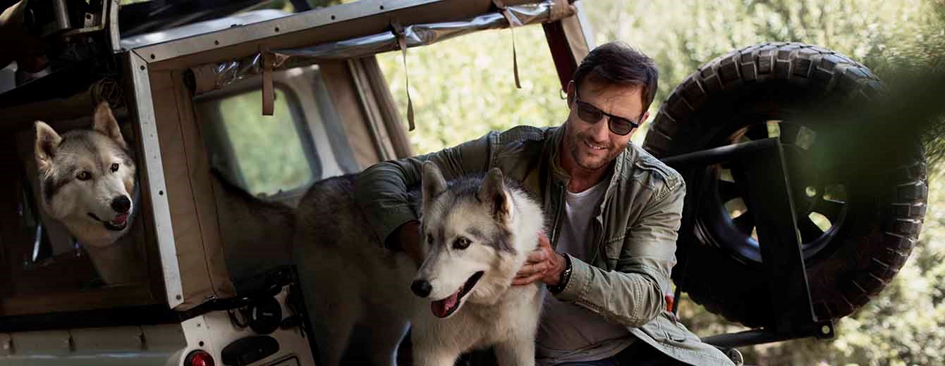 Male wearing dark lenses in back of jeep with two husky dogs