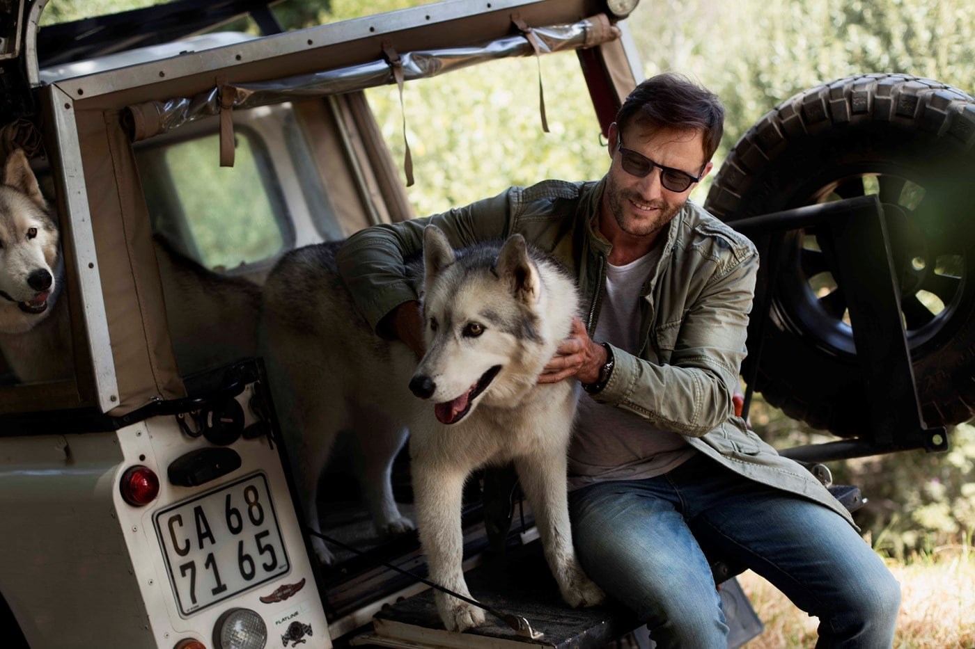 Male wearing dark lenses in back of jeep with two husky dogs